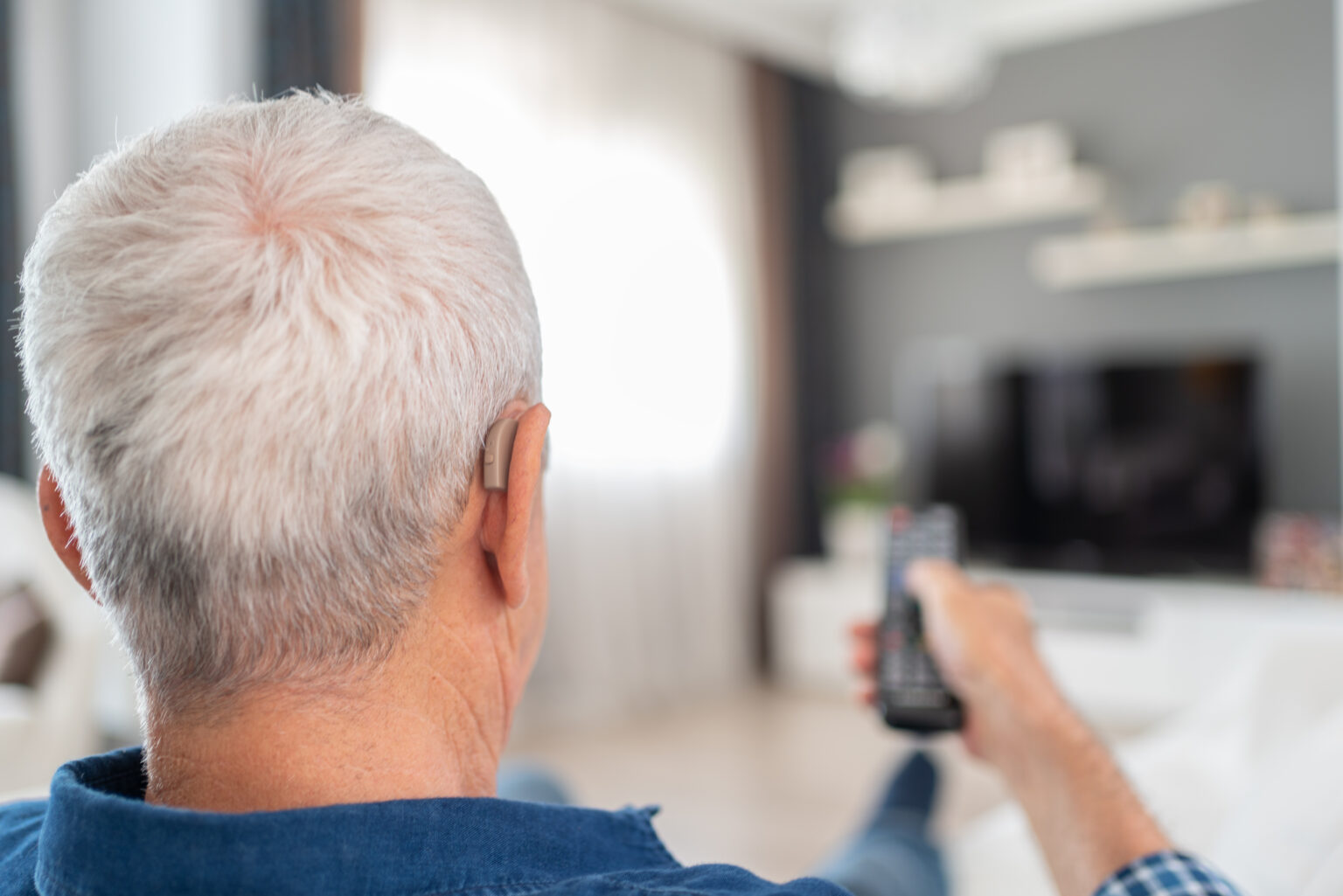 Man with hearing aids holds up the remote control to turn on the TV. He is able to comfortably watch TV because he wears hearing aids.