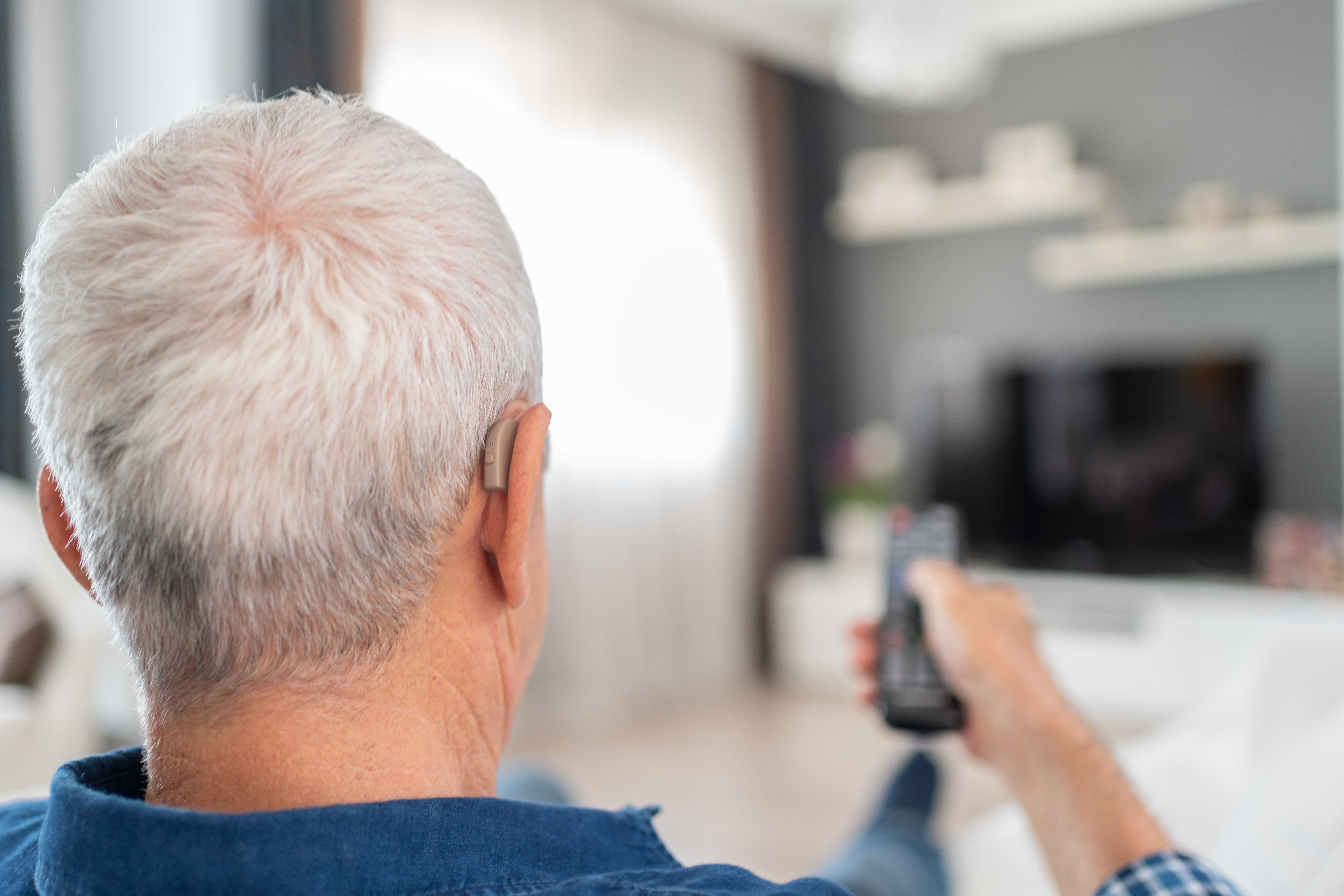 Man with hearing aids holds up the remote control to turn on the TV. He is able to comfortably watch TV because he wears hearing aids.