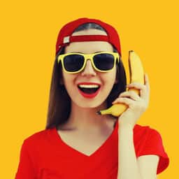 Portrait of a woman holding a banana like a phone against an orange background.