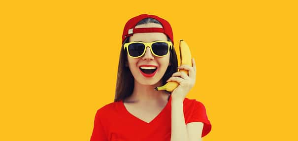 Portrait of a woman holding a banana like a phone against an orange background.
